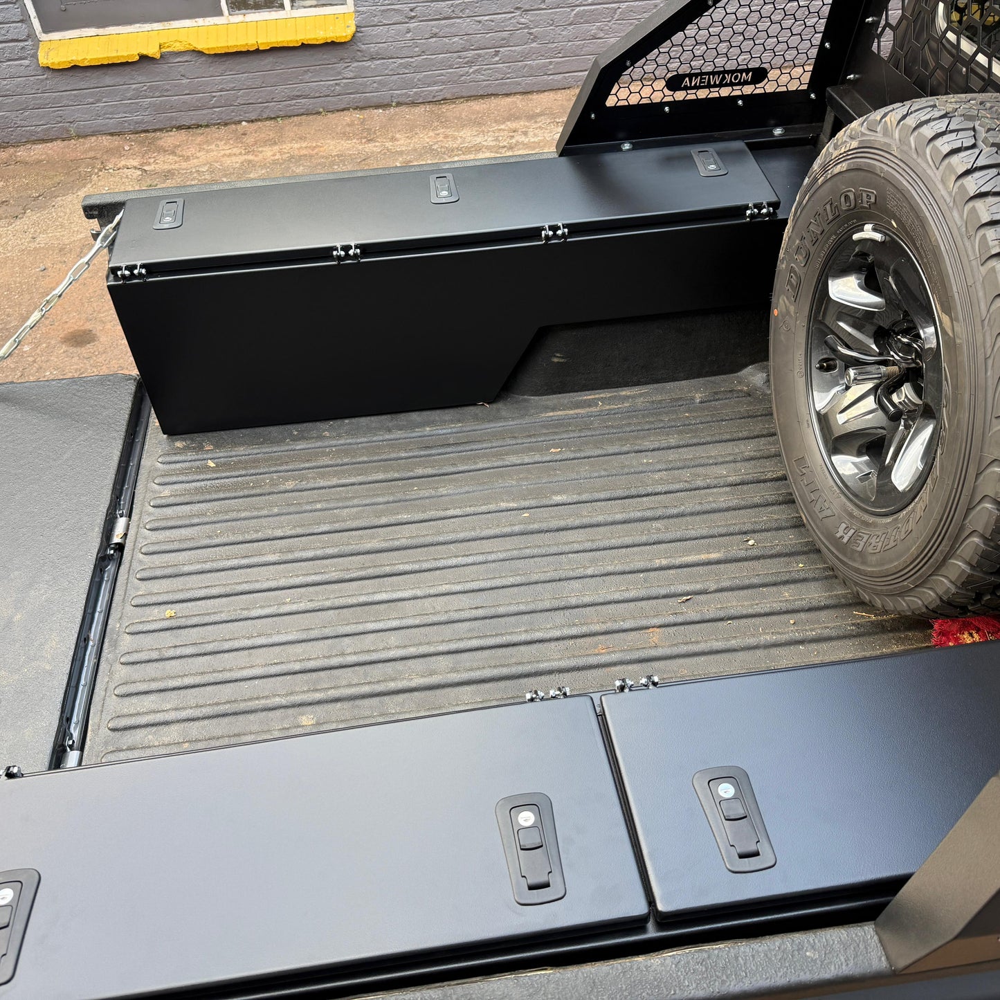 Truck bed with a black toolbox and tire on a concrete surface.