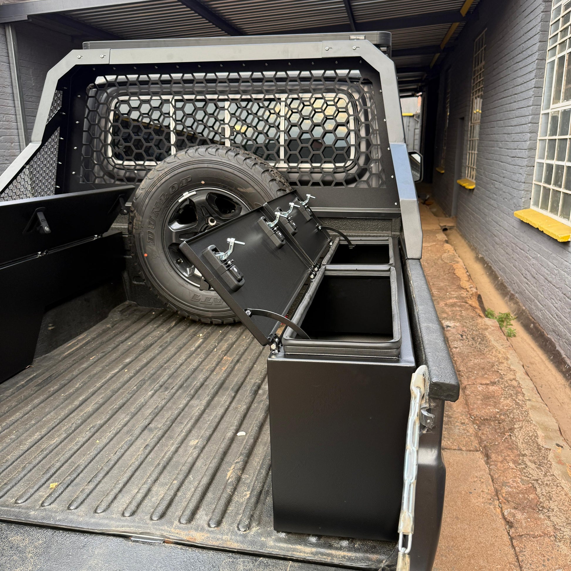 Truck bed with storage box and tire, parked next to a building.
